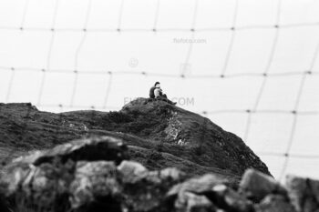 Two people sitting on a hill overlooking the sea viewed through a hole in a wire mesh fence. Swansea, Wales. 1977.