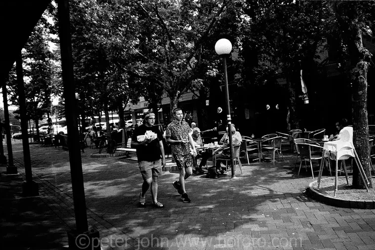 Two young men carrying drinks and snacks walking past cafes in the vibrant Corbett Plaza, Bowral, NSW, Australia.